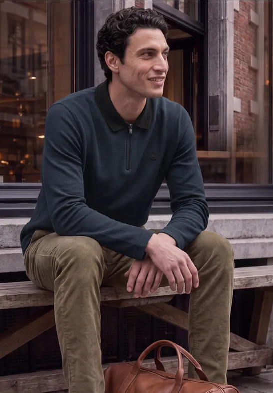 Un homme souriant est assis sur un banc en bois devant un bâtiment, avec un sac en cuir à côté.