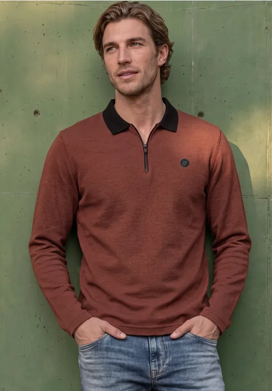 Ein lässig gekleideter Mann mit braunem Langarm-Poloshirt und Jeans vor grüner Wand.