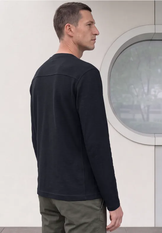 Ein Mann in schwarzem Langarmshirt steht seitlich vor einer hellen Wand mit rundem Fenster.