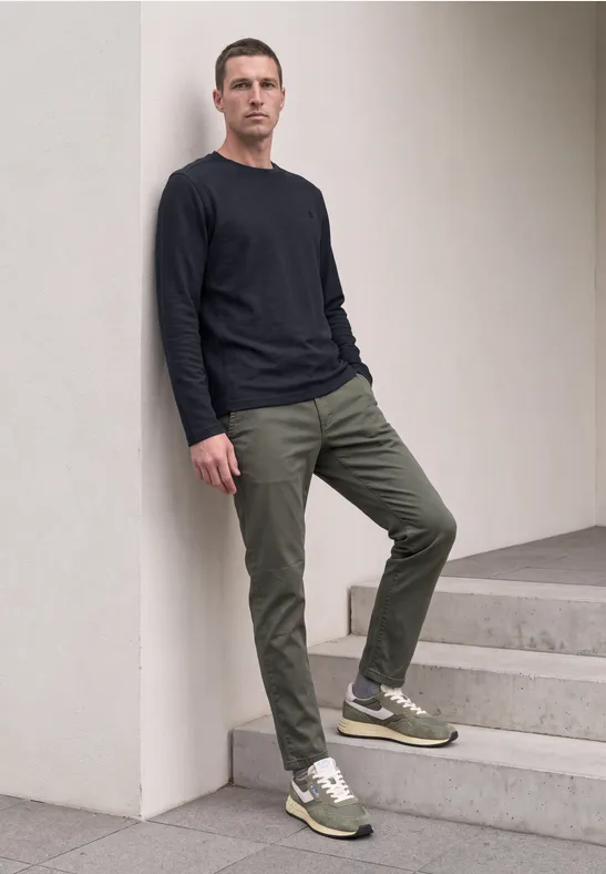 Mann lehnt an weißer Wand, trägt schwarzes Shirt, olivgrüne Hose und grüne Sneaker.