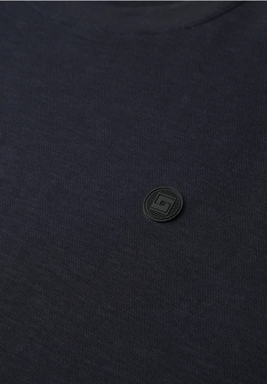 Nahaufnahme eines dunklen T-Shirts mit einem runden, dezenten Markenemblem.