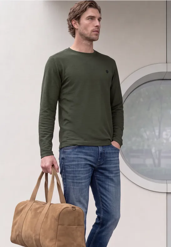 Ein Mann steht in Jeans und olivgrünem Langarmshirt, hält lässig eine braune Reisetasche.