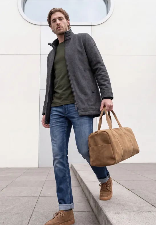Ein Mann in Jeans, grünem Shirt und grauem Mantel spaziert mit einer braunen Reisetasche.