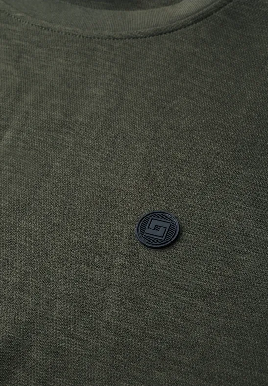 Grünes Strickshirt mit rundem schwarzem Logo-Pin auf der Brust.
