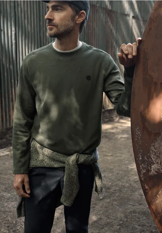 Ein Mann in grünem Sweatshirt und Cap, Pullover um die Hüfte gebunden, steht vor einer Metallstruktur im Freien.