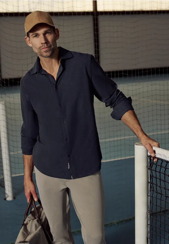 Eine Person lehnt am Tennisnetzpfosten, trägt Hemd, Hose und hält eine Tennistasche.