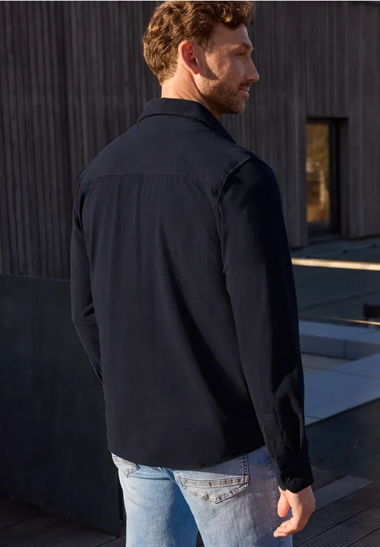 Mann steht mit dem Rücken zur Kamera, trägt schwarzen Hemd und Jeans vor modernem Betonbau.