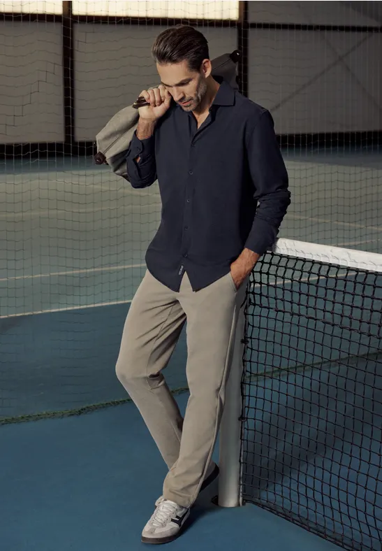 Ein Mann trägt Hemd und Hose, steht lässig mit Jacke über der Schulter an einem Tennisnetz.