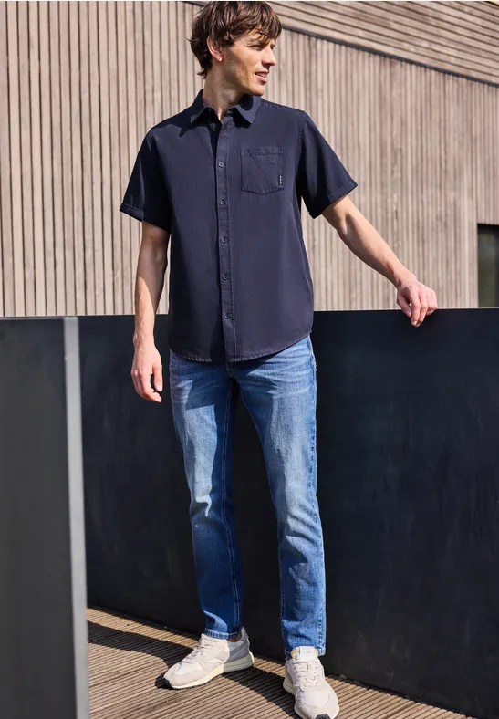 Ein Mann steht vor einer Holzfassade und trägt ein dunkles Hemd, blaue Jeans und weiße Sneaker.