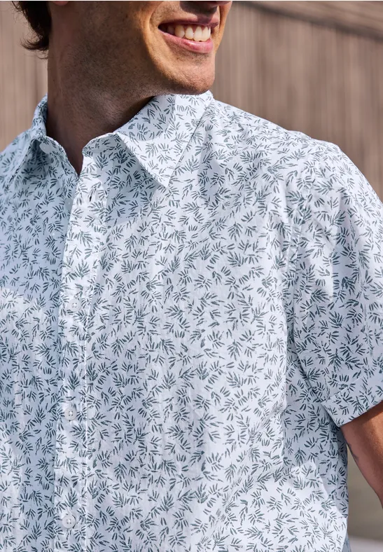 Mann in weißem Hemd mit blau-grünem Blattmuster lächelt.