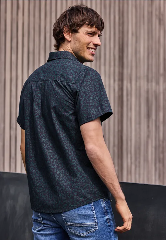 Mężczyzna tyłem w ciemnej koszuli z wzorem i jeansach, patrzy przez ramię na drewniane tło.