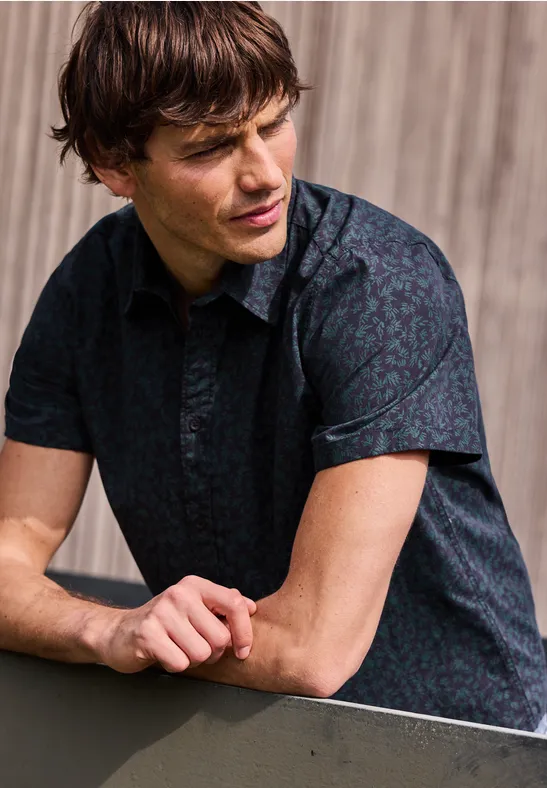 Mężczyzna opiera się o balustradę, ma na sobie ciemną, wzorzystą koszulę z krótkim rękawem na tle drewna.