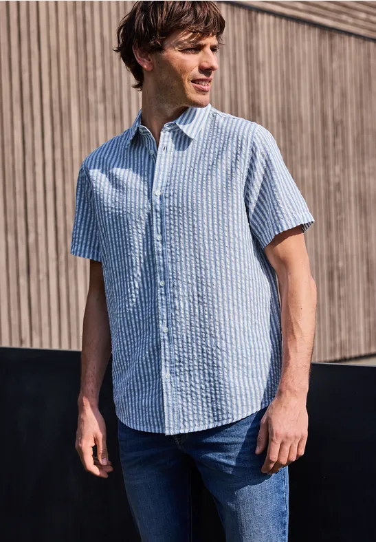 Ein Mann mit verschwommenem Gesicht trägt ein hellblaues, kurzärmliges Hemd vor einer Holzverkleidung.