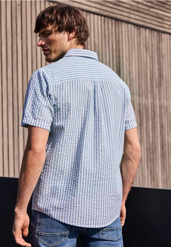 Ein Mann steht mit dem Rücken zur Kamera, trägt ein kurzärmeliges blau-weiß kariertes Hemd vor einer Holzwand.