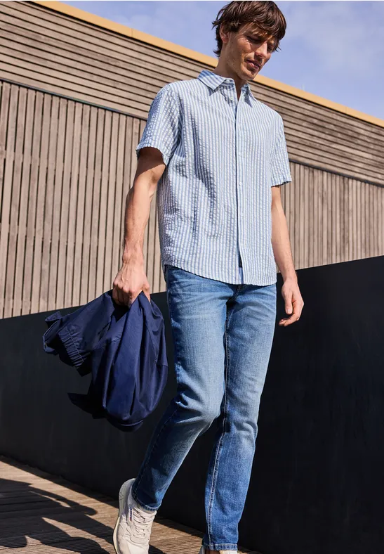 Eine Person trägt ein hellblaues Kurzarmhemd, Jeans und weiße Sneaker, hält eine Jacke und steht im Freien.