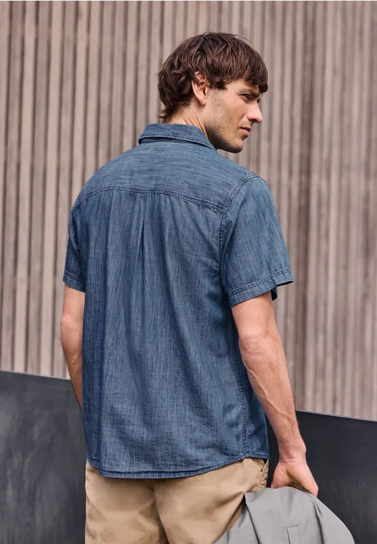 Ein Mann von hinten in kurzärmligem Jeanshemd und beigen Chinohosen vor einem vertikal strukturierten Hintergrund.