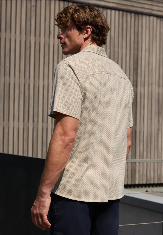 Een man staat buiten met zijn rug naar de camera voor een houten gevel, gekleed in een licht overhemd en donkere broek.