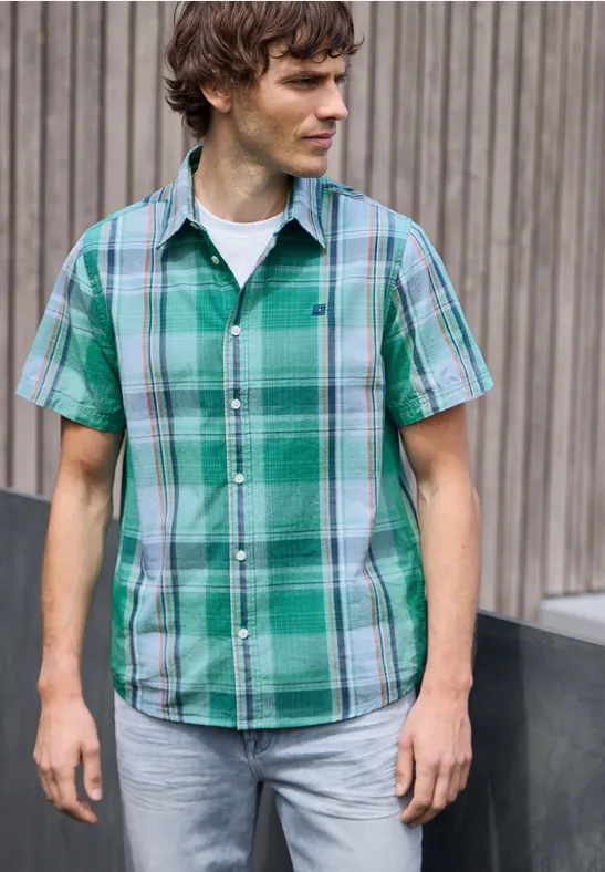 Ein Mann steht vor einer Holzfassade und trägt ein grün kariertes Kurzarmhemd und Jeans.