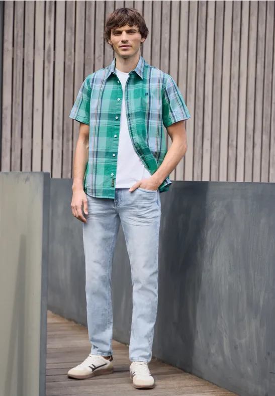 Ein junger Mann in grün kariertem Hemd und Jeans steht lässig vor einer Holzverkleidung.