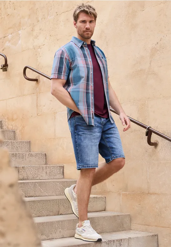 Een man in zomerse kleding staat op een stenen trap en leunt tegen de leuning voor een lichte muur.