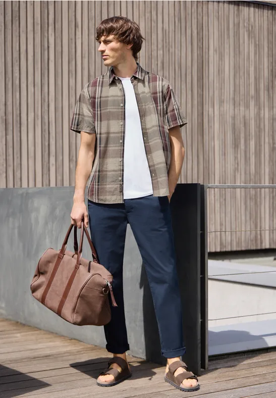 Man in geruit overhemd, t-shirt, wijde broek en sandalen met een reistas voor een houten wand.