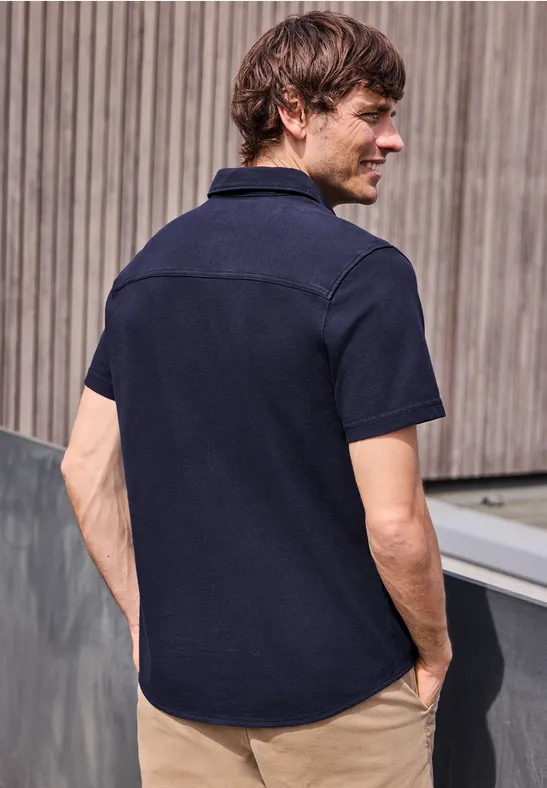 Ein Mann steht mit dem Rücken zur Kamera, trägt ein dunkelblaues Kurzarmhemd vor Holzfassade.