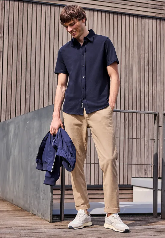 Junger Mann in navyblauem Hemd, hellen Chinos und weißen Sneakern, hält eine blaue Jacke und steht im Freien.