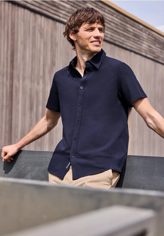 Mann in dunkelblauem Kurzarmhemd lehnt an Metallbrüstung vor einer Holzfassade.