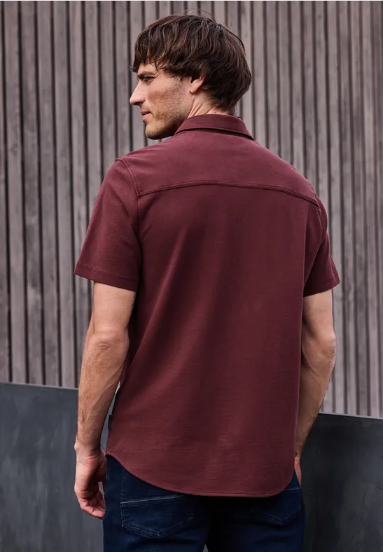 Een man, gezien van achteren, draagt een bordeaux rood overhemd en spijkerbroek voor een houten lattenwand.