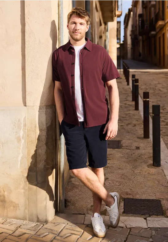 Een man leunt tegen een muur in een zonnig oud straatje, draagt een bordeaux overhemd, short en witte sneakers.