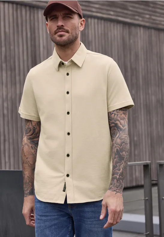 Beige overhemd met korte mouwen en knopen, tatoeages op armen, voor een houten wand.