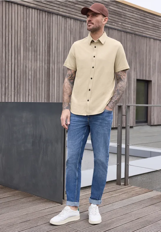 Een man draagt een lichte beige blouse, blauwe spijkerbroek en witte sneakers en leunt tegen een houten wand.