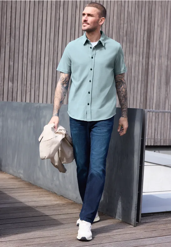 Ein Mann in hellgrünem Hemd, blauen Jeans und weißen Sneakers geht auf einem Holzdeck und hält eine Jacke.