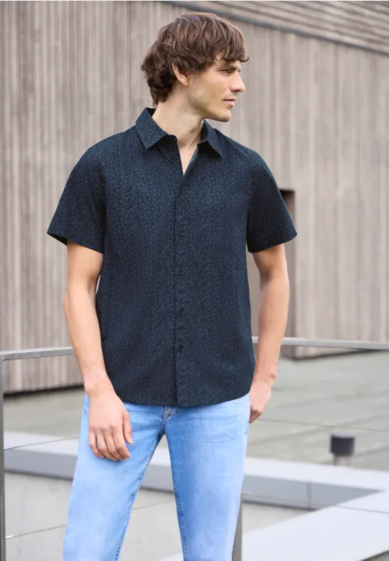 Ein Mann in dunkelblauem Kurzarmhemd und hellblauen Jeans steht vor einer Holzfassade.