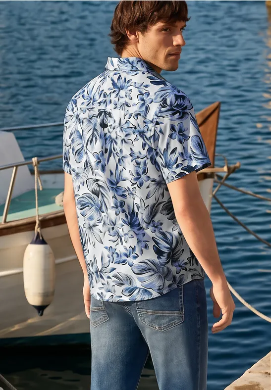 Ein Mann in blau-weiß gemustertem Hemd und Jeans steht am Bootssteg vor einem Boot auf dem Wasser.
