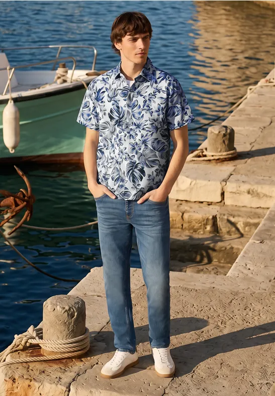 Ein Mann in floraler Kurzarmbluse und Jeans steht am Hafendamm neben einem Boot, Strohhut am Boden.