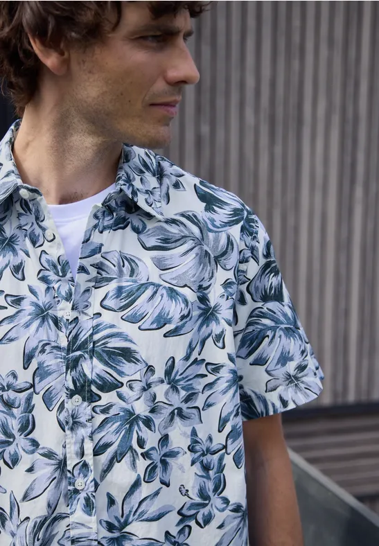 Personne au visage flouté porte une chemise à manches courtes à motif de feuilles, devant un mur vertical.