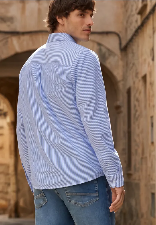 Ein Mann in blauem Hemd und Jeans steht mit dem Rücken zu uns in einem sandsteinfarbenen Gewölbegang.