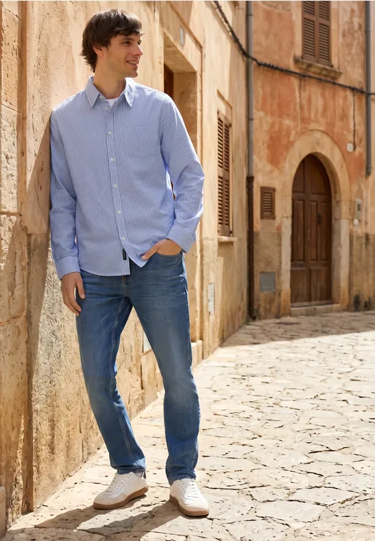 Ein Mann in hellblauem Hemd, Jeans und weißen Sneakern lehnt entspannt an einer Steinmauer in einer sonnigen Gasse.