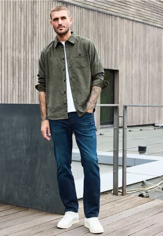 Mann in grüner Cord-Hemdjacke, weißem Shirt, blauen Jeans und weißen Sneakern auf Dachterrasse.