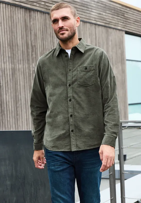 Ein Mann trägt ein grünes Cord-Hemd und blaue Jeans vor einem modernen Holzgebäude.