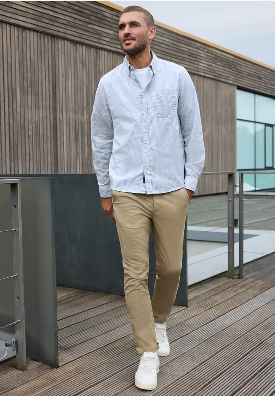 Ein junger Mann spaziert auf einem Holzsteg, trägt hellblaues Hemd, beige Hose und weiße Sneaker.