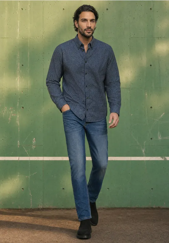 Een man in een donkerblauw hemd en jeans staat ontspannen voor een groene muur