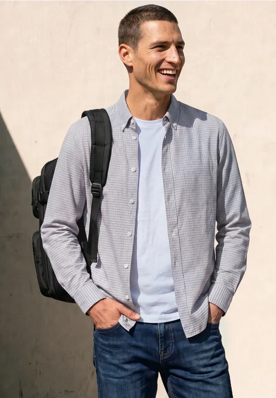 Een lachende man in jeans, wit T-shirt en lichtgrijs overhemd met zwarte rugzak voor een lichte muur.