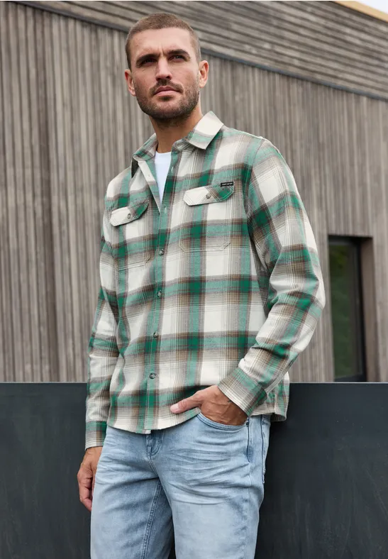 Ein Mann in kariertem Hemd und Jeans steht vor einer Holzfassade.