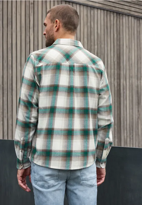 Ein Mann steht mit dem Rücken zur Kamera, trägt kariertes Hemd und Jeans vor Holzfassade.