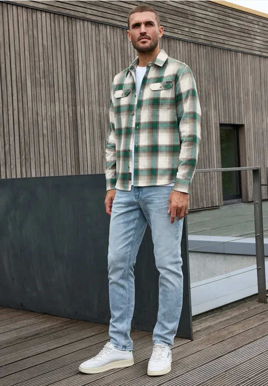 Ein Mann steht auf einer Terrasse vor Holzfassade, trägt grün-weiß kariertes Hemd, helle Jeans und weiße Sneaker.
