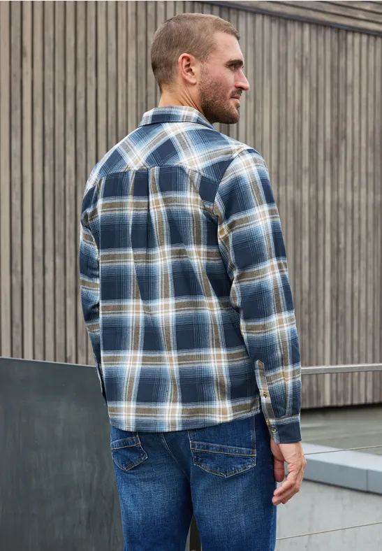 Ein Mann steht seitlich und wendet sich ab, trägt ein blau-weiß kariertes Hemd und Jeans vor einer hölzernen Wand.