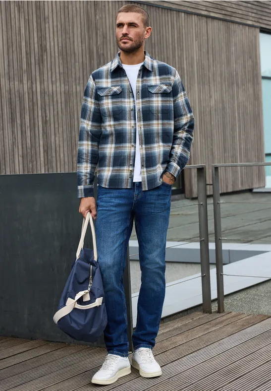 Mann in kariertem Hemd, Jeans und weißen Sneakern hält draußen eine blaue Tasche.