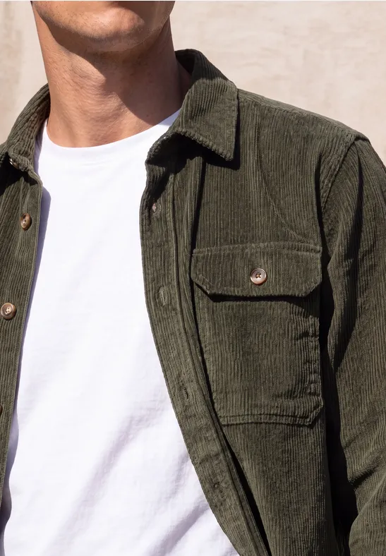 Un homme porte une veste en velours côtelé olive sur un t-shirt blanc devant un mur clair.